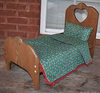Primitive wooden doll bed with green floral bedding and red lace trim, heart cutout on headboard, placed on wooden floor outdoors in front of brick wall and window.