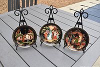 Three miniature lacquer plates displayed on metal easels on outdoor table showing intricate paintings and gold border detailing.