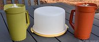 Shows two Tupperware pitchers, one green and one orange, and a white cake saver with yellow base on outdoor table, illustrating overall lot contents.