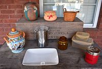 Overview of lot on wooden crate showing pottery vase, ceramic canister, stainless steel canister, white porcelain tray, plastic containers, amber glass container, red treat container and basket.