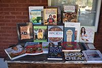 Collection of 20 assorted cookbooks displayed on a wooden surface outdoors near a brick wall. Covers include a variety of topics such as smoothies, whole30, and French wines.