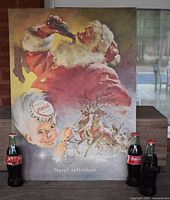 Photo showing vintage Coca-Cola Travel Refreshed poster with Santa Claus drinking Coke, character with bottle cap hat, reindeer sleigh, and three unopened 8oz classic Coca-Cola bottles in front.