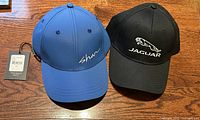 Photo of two brand new baseball caps on a wooden surface: one black Jaguar cap with white logo and text, one blue Shan cap with white script logo and original tag attached.