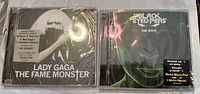 Photo showing two sealed CD jewel cases: Lady Gaga The Fame Monster on left, Black Eyed Peas The E.N.D. on right, on white background.