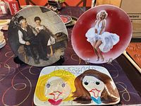 Photo shows three collectible plates: Marilyn Monroe 'Seven Year Itch' porcelain plate in front center, Norman Rockwell vintage plate to left, and colorful Cordon Bleu Girlfriends decorative plate with two female faces in front.
