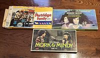 Photo showing three vintage board game boxes from the 1970s: The Partridge Family, The Hardy Boys, and Mork & Mindy, laid flat on a wooden floor.
