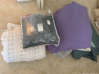 Overview of the white crocheted throw, packaged floral printed item, purple wool blanket, and additional fabrics on carpet