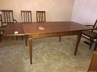 Rectangular wooden dining table with one side leaf extended, tapered legs, and decorative detail on legs. Four matching chairs in the background.