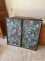 Two framed fabric art pieces leaning against a wall on carpeted floor. Both feature patchwork fabric designs in blue, green, and brown tones. Frames show light wear on edges and have no glass.