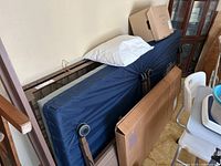 Side view of disassembled hospital bed frame stacked with navy blue mattress and white pillow on top, a cardboard box is placed at rear