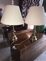 Two matching table lamps with polished brass metal bases and off-white pleated fabric lampshades placed on a wooden surface.