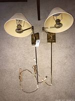 Pair of brass wall lamps with adjustable arms and cream fabric shades laid on carpet, showing cords and rectangular backplates.