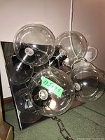 Top view of the light fixture showing four clear glass globe bulb holders attached to a square metal backplate with a reflective surface. A green sticker indicates dimensions.