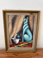 Full view of framed oil painting showing blue cat sculpture, black jug, white bowl on multicolor cloth, wooden frame with wear