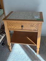 Front-facing photo showing the wood side table with a stone inset top, a front drawer with a keyhole and key, and a lower shelf. The table rests on four turned legs.