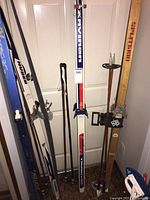 Four pairs of cross country skis standing against a door, various brands include Karphite Diamond Grind 200, Huber Touring, Splitkein with wood finish, and Jarvinen skis with red, white, and blue colors. Some skis have bindings attached.