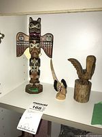Photo of the lot on display shelf showing the wood totem sculpture signed by Patrick Sealer and two smaller carved bird sculptures, all carved and painted wood.