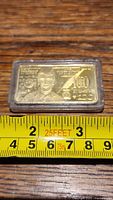 Front view of the Donald Trump $100 commemorative bar inside a plastic protective case with measuring tape for size reference.