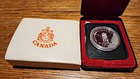 Silver dollar coin in protective capsule inside red velvet and black presentation case with white outer box bearing Canadian coat of arms.