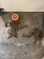 Orange 1 ton capacity chain hoist with rusted chains and hooks on concrete floor.