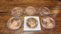 Six 1 ounce pure copper coins featuring Liberty and skull designs, displayed on wooden surface in holders.