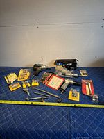 Full spread of air compressor tools and accessories on a blue padded surface with a measuring tape for scale. Includes air wrench, drill, various packaged accessories, and loose fittings.