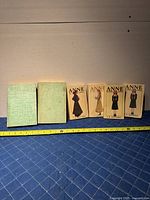 Photo showing two hardcover books with green cloth covers and four paperback books with illustrated covers of Anne of Green Gables characters, arranged in a row on a blue surface with measuring tape in front.