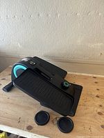 Cubii elliptical workout machine with black and turquoise design, positioned on a wooden surface with two black foot pads in front.