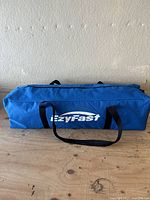 Blue rectangular carry bag with EzyFast branding on top and black carry straps placed on a wooden surface, wall background.