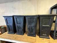 Four black resin tapered square flower planters shown from front, placed side by side on wooden shelf.