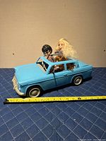Side view of blue Weasley car with three dolls seated inside and tape measure for scale.
