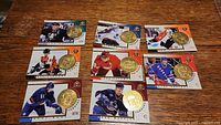 Wide view of seven hockey cards on a wooden surface, each with a gold-colored coin placed on top.
