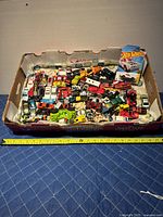 Top-down view of large box containing approx. 50 toy cars and trucks, various brands, mostly loose with some wear visible.