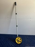 Full view of the surveyor's wheel showing the entire tool including yellow wheel and aluminum handle with black grips.