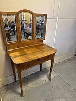Wooden vanity desk against wall showing tri-fold mirrors and single drawer.