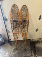 Full view of the pair of vintage wooden snowshoes leaning against a wall, showing shape, size, and materials.