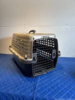 Side and front angled view of beige and black plastic pet crate on blue surface showing ventilation holes and a black metal door with locks.
