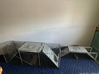 Photo showing both large and small wire mesh live animal traps placed side by side on a blue quilted surface, metal funnel entry visible on larger trap.