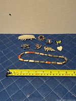 Photo showing the full lot of six brooches and one necklace arranged on a blue quilted surface with measuring tape for scale.