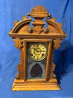 Front view of antique wooden gingerbread mantel clock showing ornate carved frame and glass front with etched designs. Roman numeral clock face visible inside the glass door.
