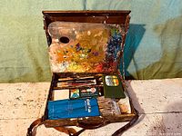 Artist's box opened showing inside contents including paint palette fixed to lid, brushes, pencils and folders of supplies