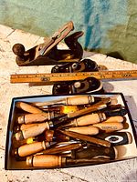 Photo showing three woodworking planes, one Stanley and two smaller Makita planes, placed behind a box full of wooden-handled chisels.