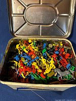 Open view of metal picnic tin loaded with colorful plastic toy cowboys and soldiers, including horses.