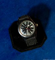 Vintage 1950s Wolbrook men's wristwatch on black display stand against blue background, angled view showing black dial and bezel.