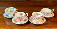 Four pairs of fine bone china tea cups and matching saucers arranged on a wooden surface. Each cup and saucer displays different floral patterns with gold rim accents, featuring roses, mixed flowers, and green leaves.