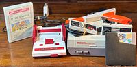 Wide shot showing gray NES console on right with two controllers and orange Zapper, red and white Famicom console on left, Micro Genius cartridge in case and adapter in foreground.