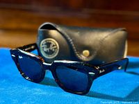 Black Ray-Ban Wayfarer sunglasses displayed in front of original black hard leather case with Ray-Ban logo. Sunglasses show classic black trapezoidal frames with dark tinted lenses.
