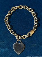 Full view of the sterling silver rolo link bracelet with attached heart charm laid flat on a dark cloth background.
