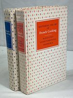 Two hardcover books standing side by side showing white dust jackets with red and blue fleur-de-lis patterned design and labeled with Mastering the Art of French Cooking