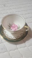 Aynsley bone china teacup on matching saucer showing pink rose floral design and gold trim on white base with dark green and gold filigree on saucer.
